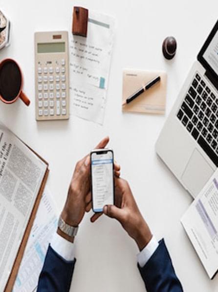 person holding phone with calculator and computer at desk